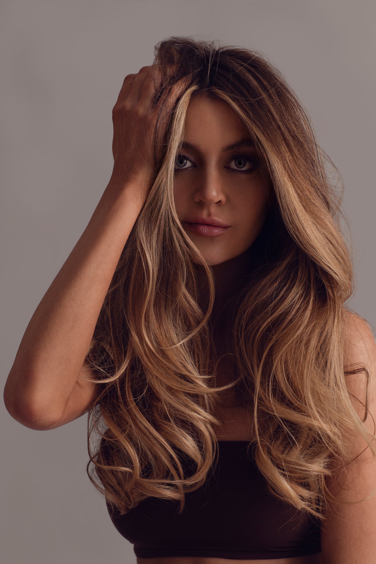 A woman with long, voluminous, wavy blonde hair poses against a neutral background in a brown top, touching her hair while looking at the camera, enhanced by Oriza rice water infused hair care