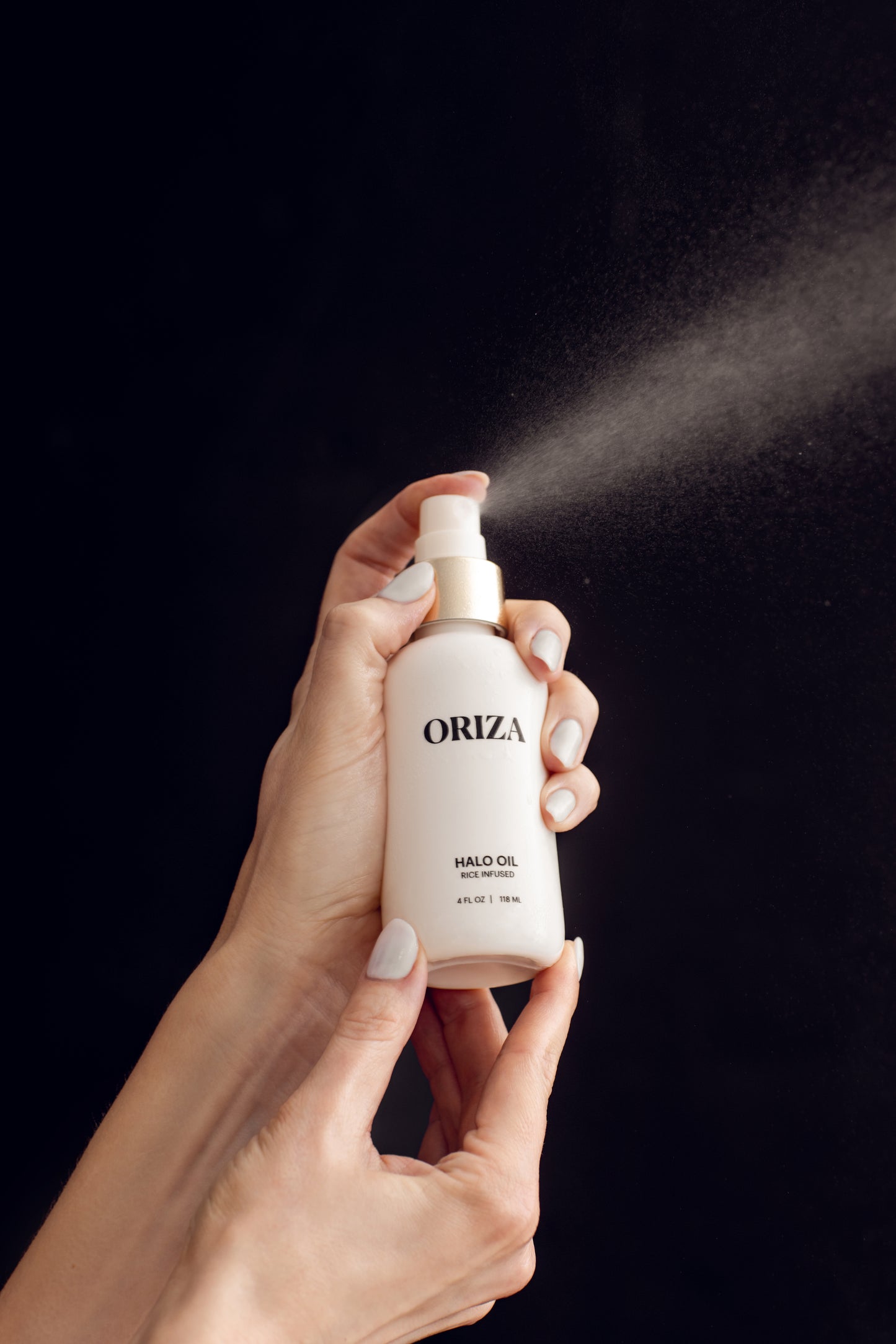 Hands hold a white bottle of Oriza Halo Oil Mist as a fine spray releases, capturing its lightweight texture.