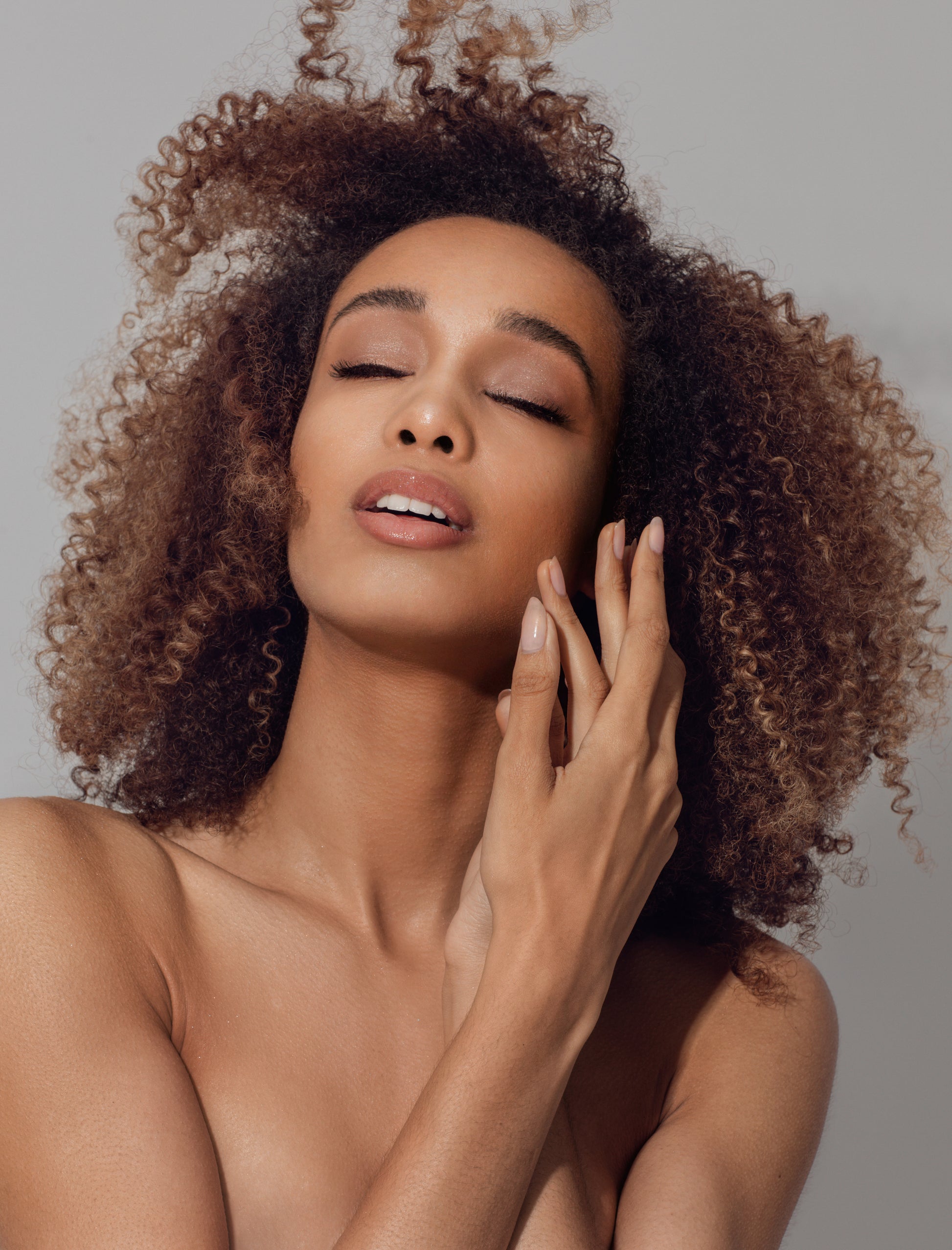 A woman with naturally curly hair and glowing skin closes her eyes in a serene moment, showcasing soft, hydrated curls enhanced with Oriza Halo Oil’s lightweight formula.