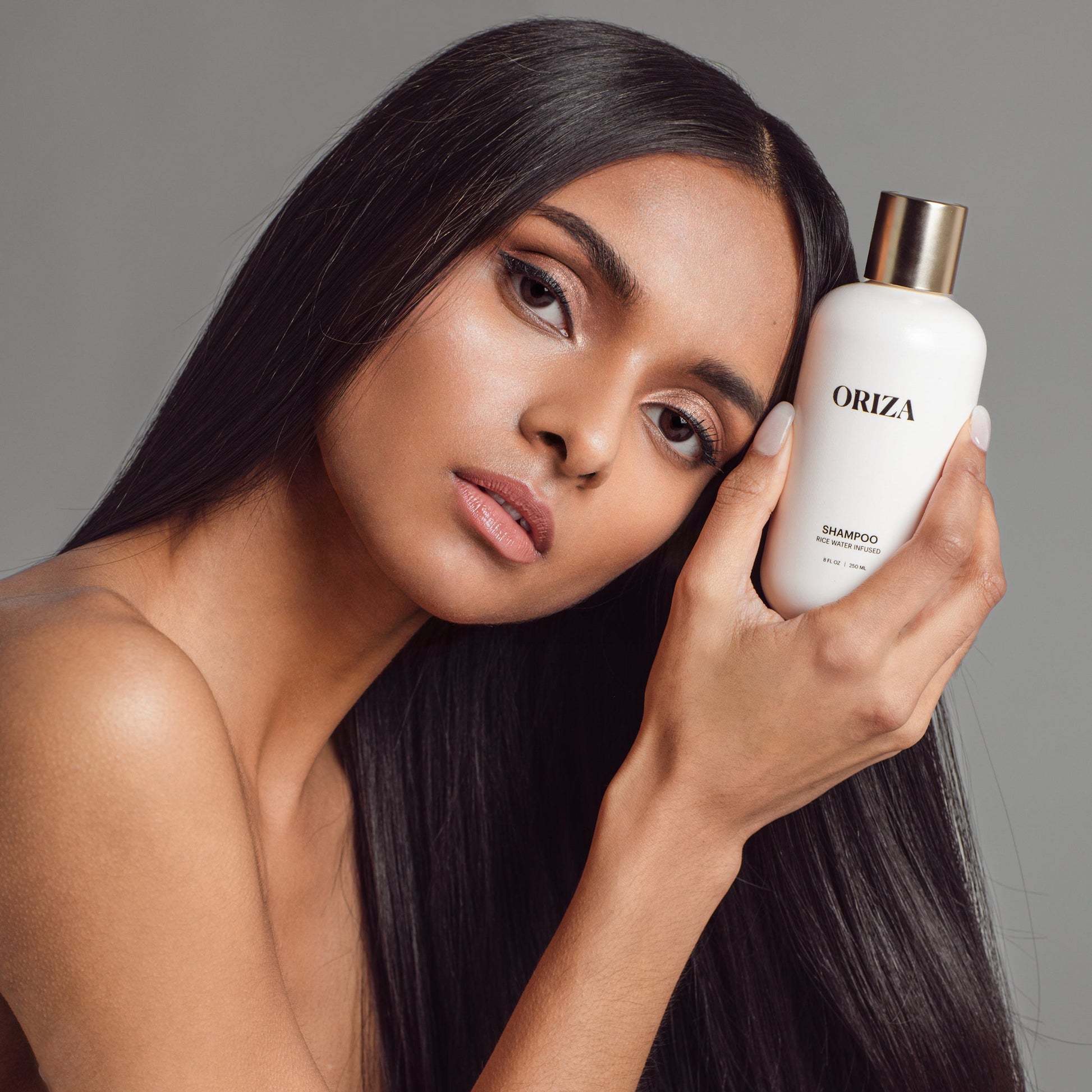 A woman with thick, long, straight black hair poses against a neutral background, holding a bottle of Oriza rice water infused shampoo next to her face.
