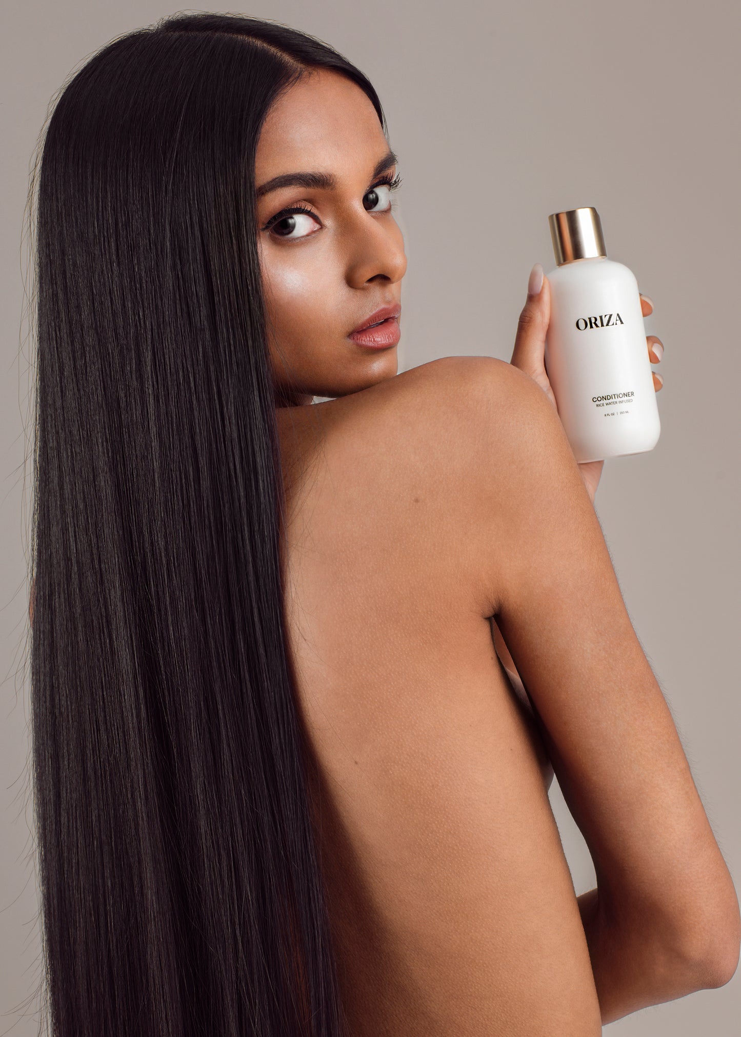 A woman with thick, long, straight black hair looks over her shoulder while holding a white Oriza rice water infused conditioner bottle with a gold cap against a neutral background.