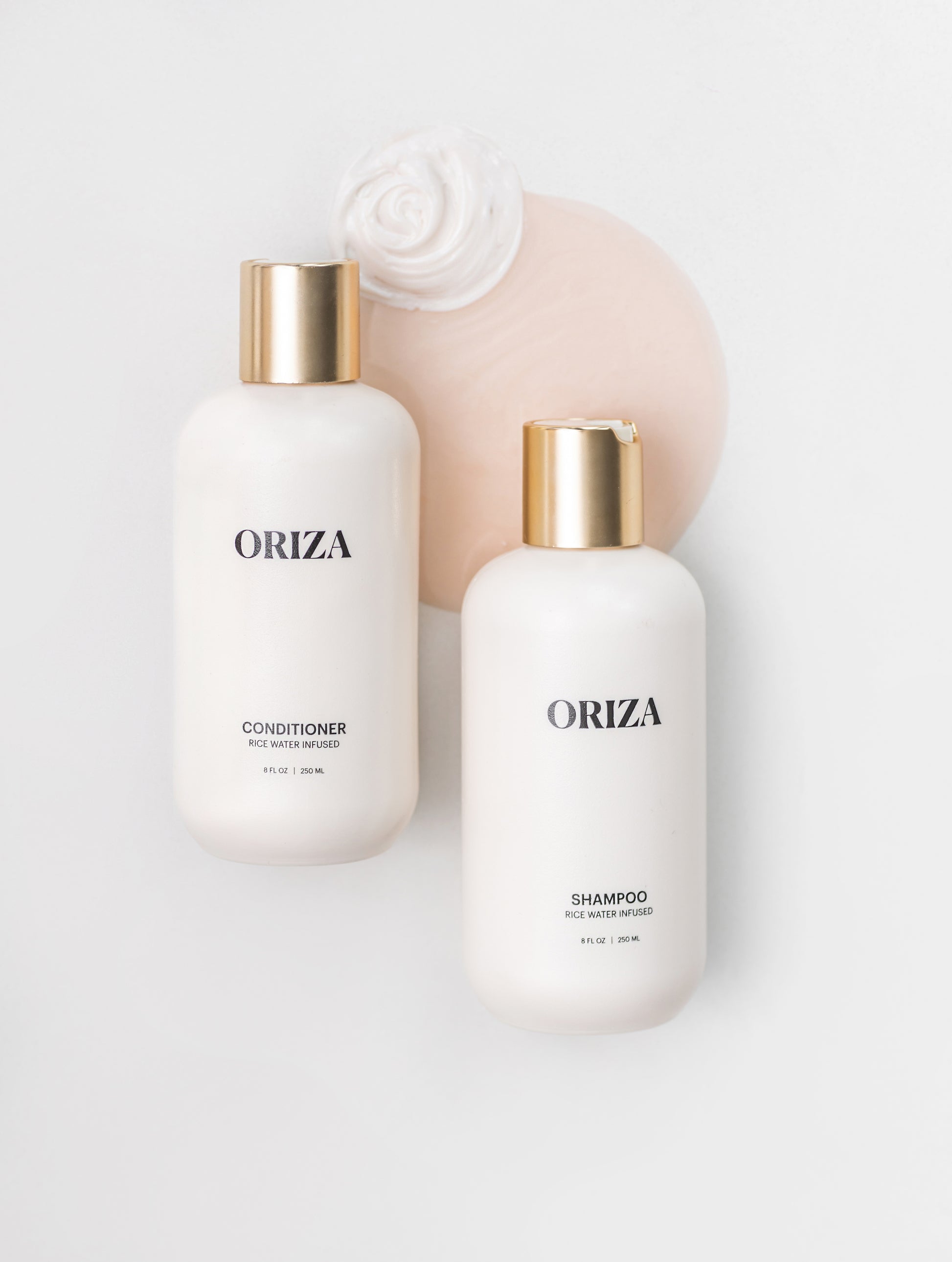 Two white bottles of Oriza rice water infused shampoo and conditioner with gold caps rest on a light surface beside swirls of creamy product, highlighting their luxurious, nourishing formula.
