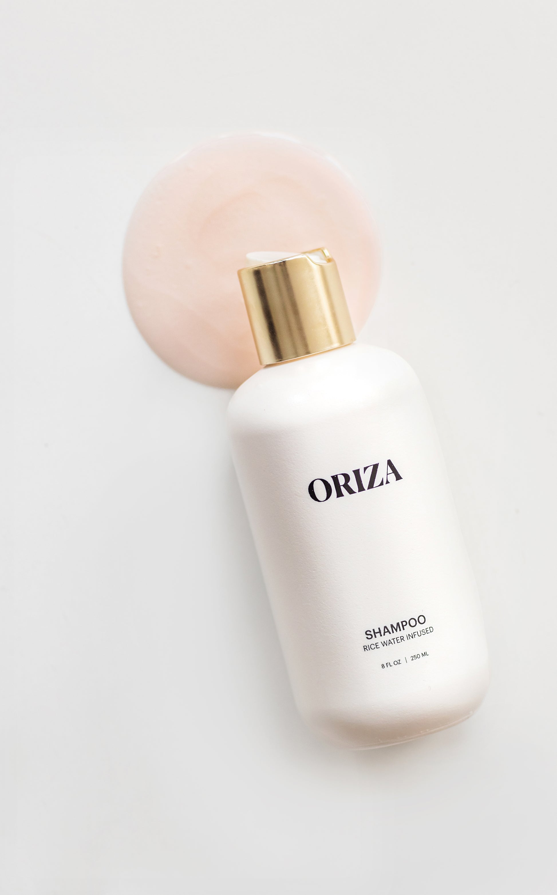 A white bottle of Oriza rice water infused shampoo with a gold cap pours its luxurious formula onto a clean, light background.